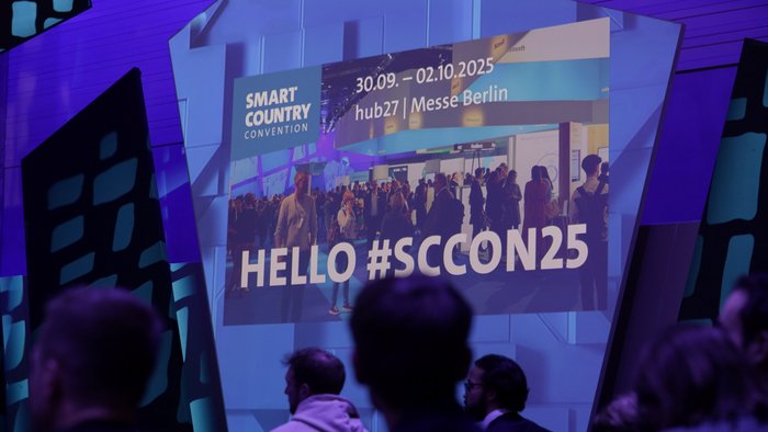 Blick auf einen großen Monitor, auf dem folgender Text zu lesen ist: "Smart Country Convention. 30.09. - 02.10.2025 hub27 | Messe Berlin. HELLO #SCCON25. 