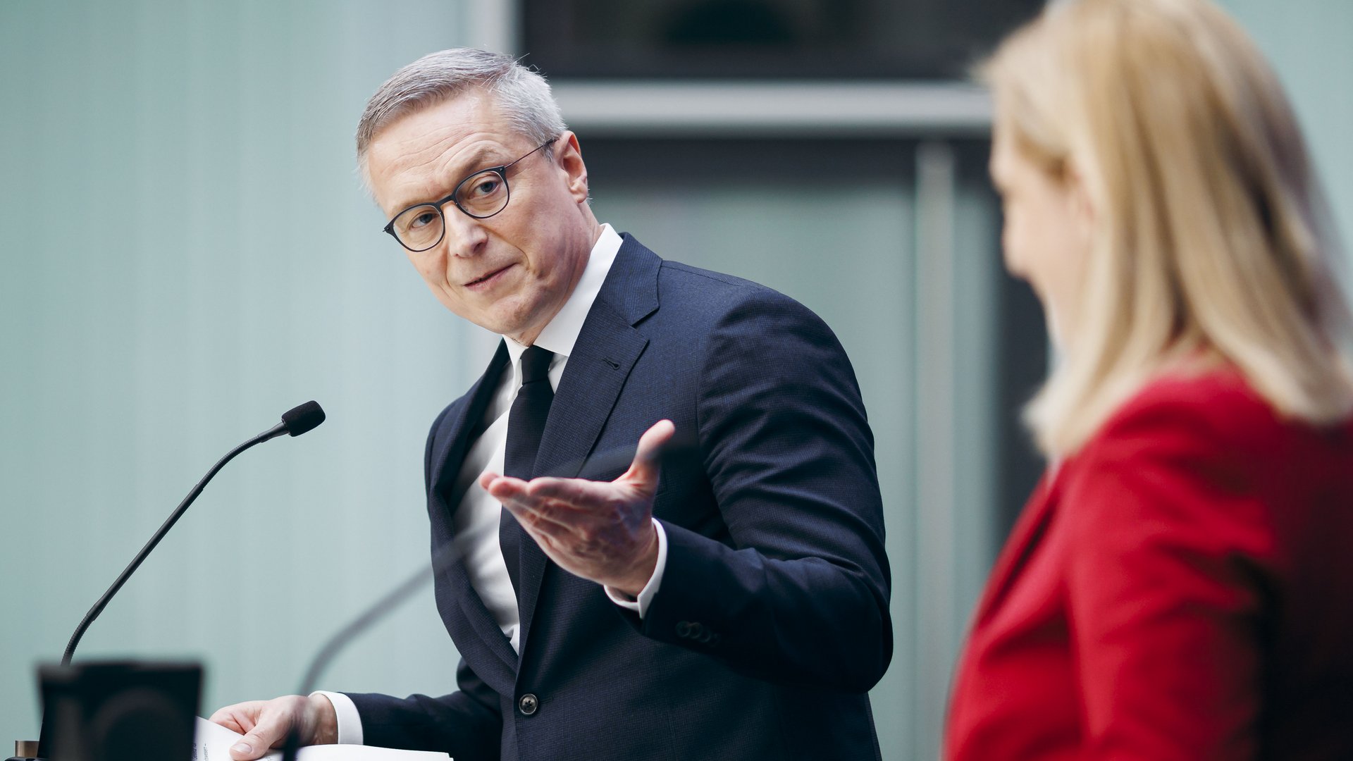 Karsten Wildberger im Anzug am Rednerpult zeigt mit der offenen Handfläche auf eine Frau, die rechts im Bild verschwommen zu sehen ist. 
