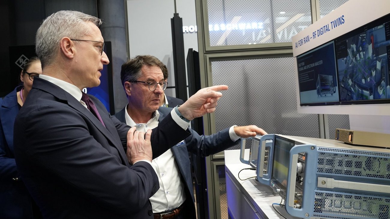 Zwei Männer im Geschäftsanzug stehen an einem Messestand von Rohde & Schwarz und begutachten eine technische Präsentation auf einem Monitor. Ein Mann zeigt erklärend auf das Display, auf dem der Text „AI FOR RAN – RF DIGITAL TWINS“ zu sehen ist. Vor ihnen auf dem Tisch stehen professionelle, hellblaue Hochfrequenz-Messgeräte.