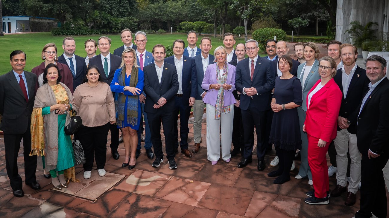 Eine Gruppe von Menschen steht am Rande einen Hauses vor einer grünen Wiese für ein Gruppenbild bereit. 