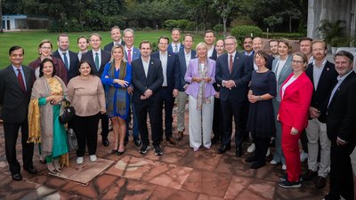 Eine Gruppe von Menschen steht am Rande einen Hauses vor einer grünen Wiese für ein Gruppenbild bereit. 
