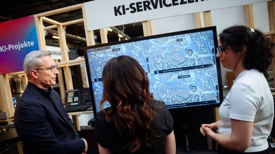 Drei Personen stehen vor einem Monitor an einem Messestand. Über dem Monitor steht der Schriftzug "KI-Servicezentrum".