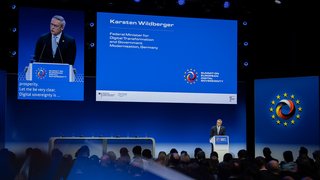 Fernaufnahme von Digitalminister Wildberger auf der Bühne mit Publikum. 
