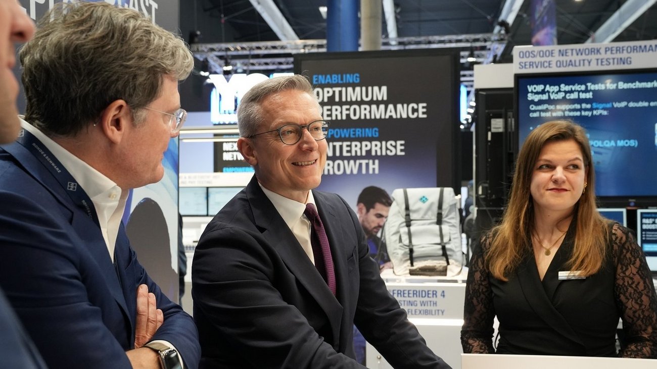 Bundesdigitalminister Dr. Karsten Wildberger (Mitte) im Gespräch mit zwei Personen an einem Messestand auf dem MWC 2026. Wildberger trägt einen dunklen Anzug und eine weinrote Krawatte und blickt lächelnd nach links. Im Hintergrund sind Monitore mit Grafiken zu „Optimum Performance“ und technisches Equipment zu sehen.