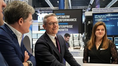 Bundesdigitalminister Dr. Karsten Wildberger (Mitte) im Gespräch mit zwei Personen an einem Messestand auf dem MWC 2026. Wildberger trägt einen dunklen Anzug und eine weinrote Krawatte und blickt lächelnd nach links. Im Hintergrund sind Monitore mit Grafiken zu „Optimum Performance“ und technisches Equipment zu sehen.