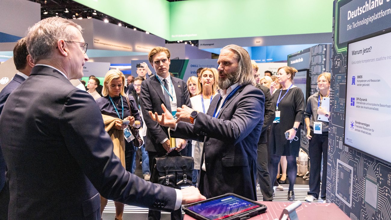 Situation an einen Messestand: eine Person im Anzug zeigt einer Delegation von Menschen etwas an einer Messewand mit Screen. Oben steht Deutschland-Stack. 