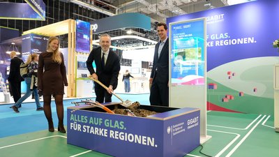 Drei Personen an einem Messestand: einer hält einen Spaten in eine kleine Baustelle, aus der Glasfaserkabel herausschauen. 