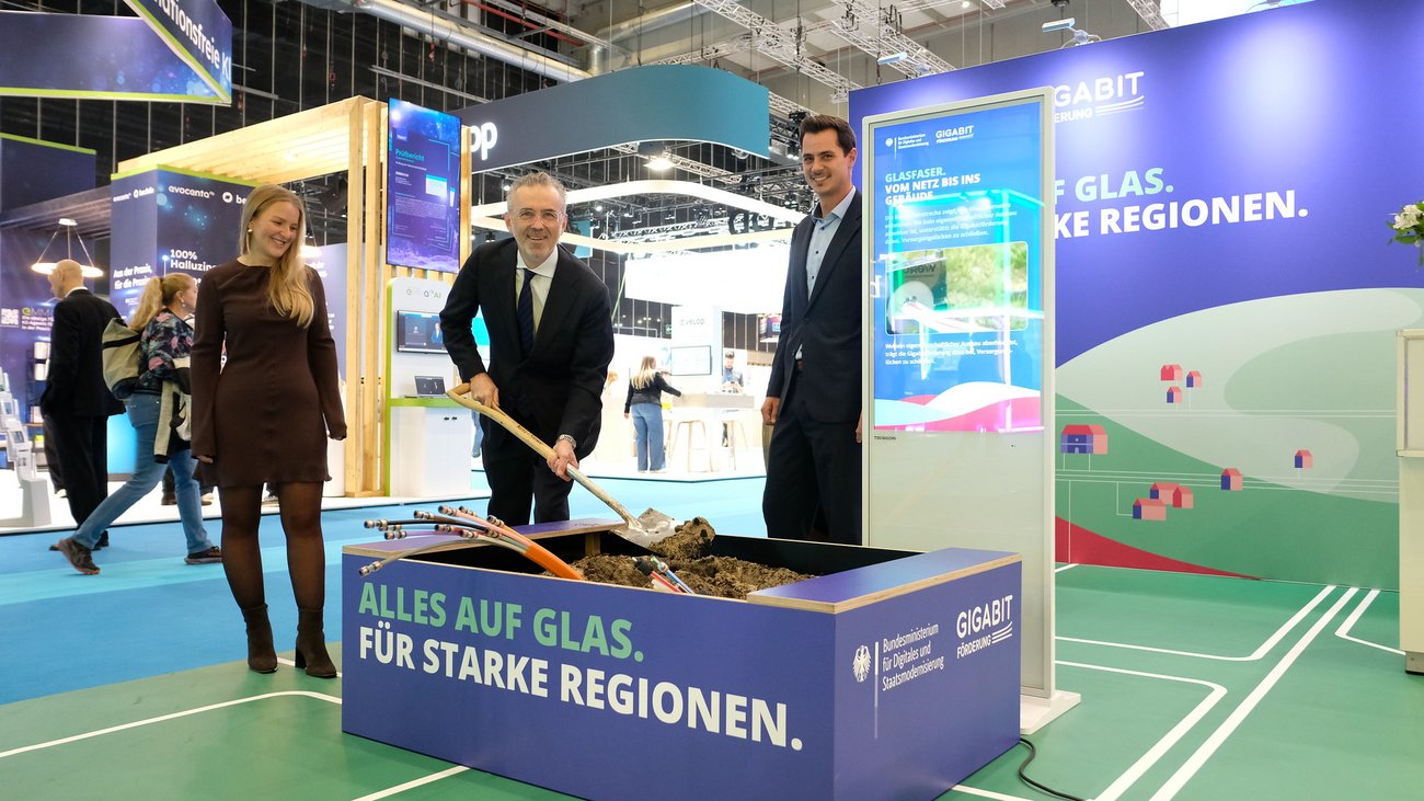 Drei Personen an einem Messestand: einer hält einen Spaten in eine kleine Baustelle, aus der Glasfaserkabel herausschauen. 