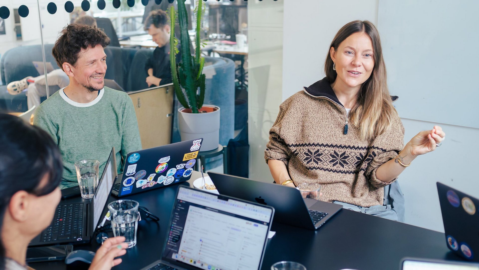 Drei Personen sitzen an einem Tisch und sprechen miteinander. Vor ihnen stehen geöffnete Laptops. Eine lächelnde Frau rechts im Bild spricht und gestikuliert mit einem Arm. Die anderen Personen hören ihr zu.