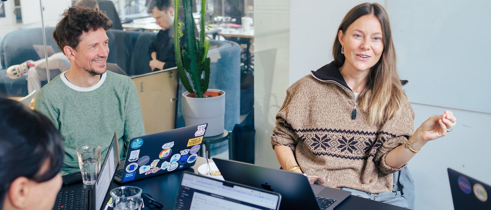 Drei Personen sitzen an einem Tisch und sprechen miteinander. Vor ihnen stehen geöffnete Laptops. Eine lächelnde Frau rechts im Bild spricht und gestikuliert mit einem Arm. Die anderen Personen hören ihr zu.