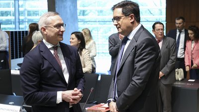 Zwei Männer im Anzug unterhalten sich stehend bei einer internationalen Konferenz; im Hintergrund sind weitere Teilnehmer sowie ein Schild mit der Aufschrift „MAGYARORSZÁG“ (Ungarn) zu sehen.