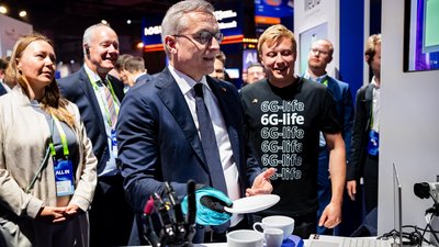 Situation an einem Messestand: ein Herr im Anzug testet neueste Technik mit einem Handschuh vor einem Laptop. Dahinter ein Mann im Shirt, auf dem "6G-life" steht. Im Hintergrund Publikum und Messestände. 