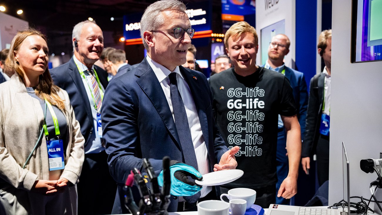 Situation an einem Messestand: ein Herr im Anzug testet neueste Technik mit einem Handschuh vor einem Laptop. Dahinter ein Mann im Shirt, auf dem "6G-life" steht. Im Hintergrund Publikum und Messestände. 