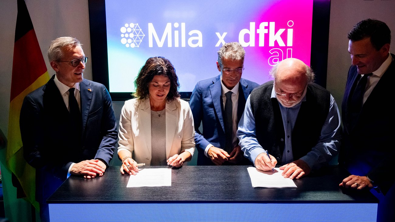 Fünf Personen am Tresen, zwei unterzeichnen Dokumente. Im Hintergrund leuchtet der Schriftzug "Mila x DFKI ai"