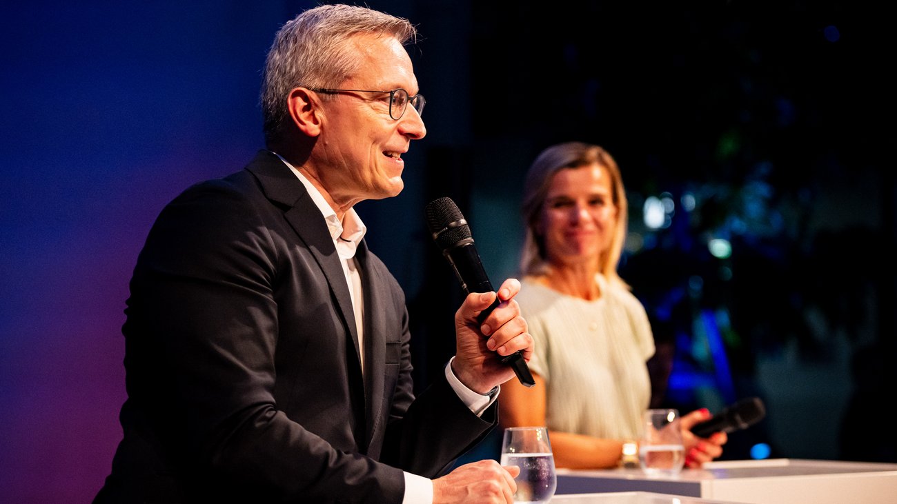 Digitalminister Wildberger spricht in ein Mikrofon, im Hintergrund verschwommen Betty Kieß.