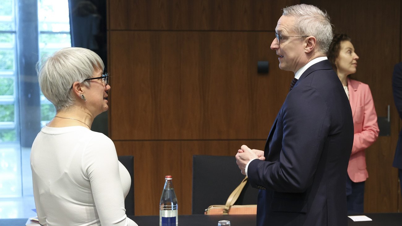 Andrea Polereczki, ungarische Staatssekretärin für Cybersicherheit mit kurzen, grauen Haaren und Brille, steht im Profil und führt ein Gespräch mit Minister Karsten Wildberger. Das Treffen findet in einem modernen Konferenzraum mit einer Holzvertäfelung im Hintergrund statt. Vor ihnen steht ein Tisch mit einer Wasserflasche, einem Glas und einem Namensschild, auf dem das Wort „MAGYAR“ (Ungarn) zu sehen ist. Im Hintergrund steht eine weitere Frau im rosa Blazer.