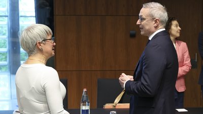 Andrea Polereczki, ungarische Staatssekretärin für Cybersicherheit mit kurzen, grauen Haaren und Brille, steht im Profil und führt ein Gespräch mit Minister Karsten Wildberger. Das Treffen findet in einem modernen Konferenzraum mit einer Holzvertäfelung im Hintergrund statt. Vor ihnen steht ein Tisch mit einer Wasserflasche, einem Glas und einem Namensschild, auf dem das Wort „MAGYAR“ (Ungarn) zu sehen ist. Im Hintergrund steht eine weitere Frau im rosa Blazer.