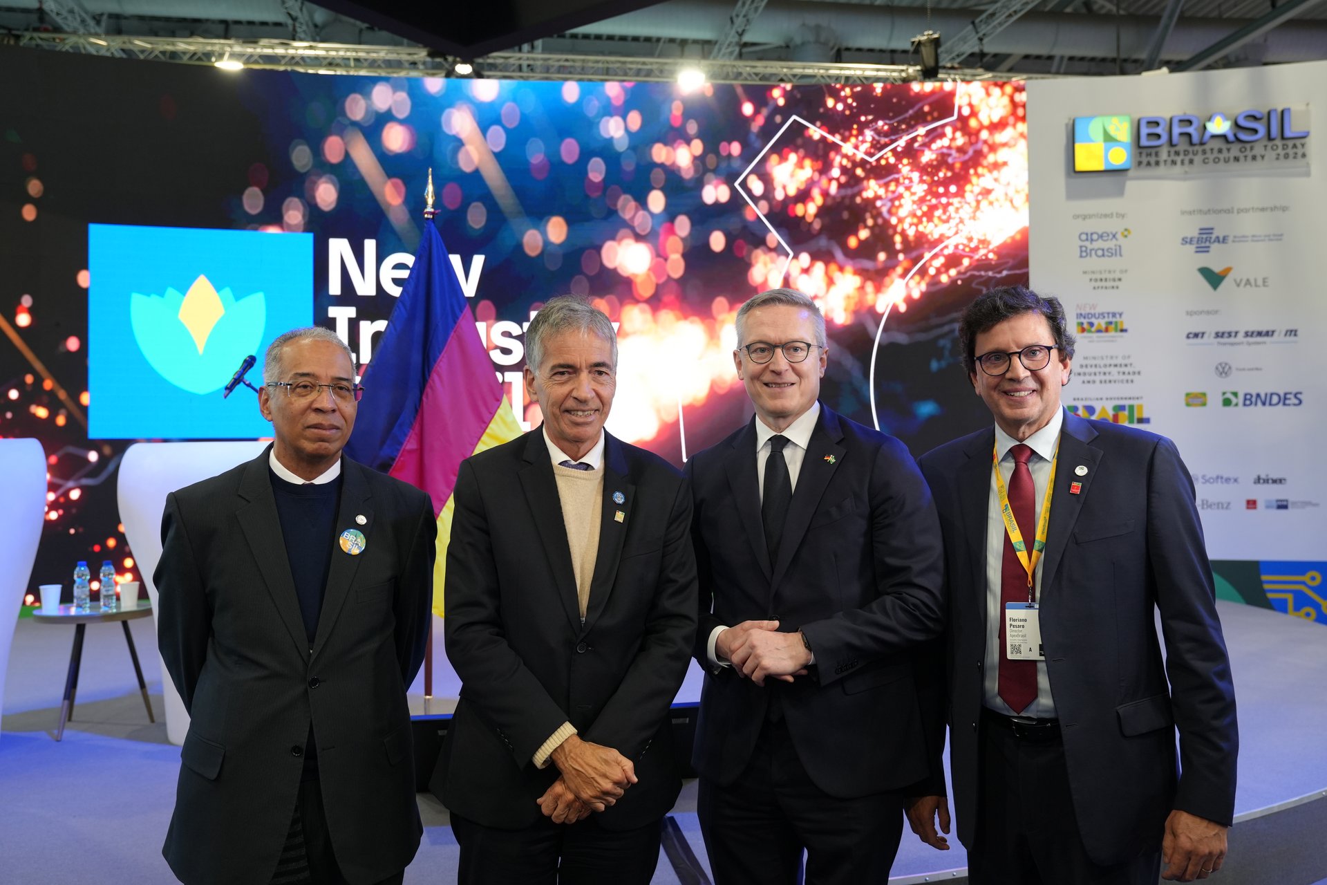 Gruppenbild von vier Männern in formeller Kleidung (Anzüge und Sakkos) auf der Hannover Messe 2026. Sie stehen lächelnd nebeneinander vor einer Veranstaltungsbühne. Im Hintergrund ist eine große Leinwand mit dem Slogan „Brasil – The Industry Partner Country 2026“ sowie Logos von Partnern wie ApexBrasil und BNDES zu sehen. Zwischen den Männern ist dezent eine deutsche Flagge platziert. Die Szene wirkt professionell und unterstreicht die deutsch-brasilianische Wirtschaftskooperation.