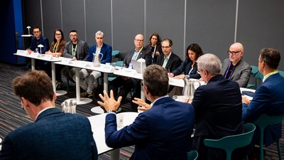 Situation in einem Sitzungsraum: viele Menschen in Businesskleidung an Tischen im U-Format.  