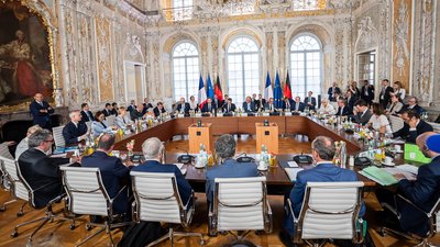 Teilnehmerinnen und Teilnehmer des Deutsch-Französischen Ministerrats sitzen an einem großen U-förmigen Konferenztisch in einem prunkvollen Saal.