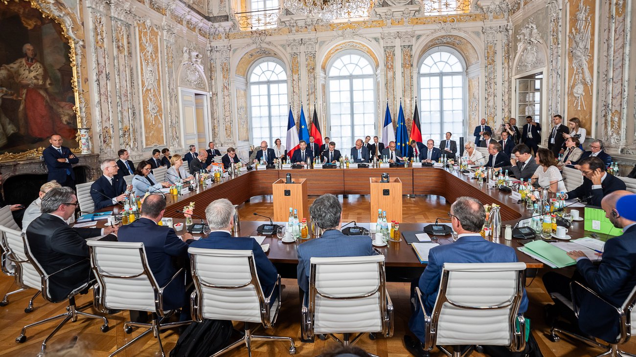 Teilnehmerinnen und Teilnehmer des Deutsch-Französischen Ministerrats sitzen an einem großen U-förmigen Konferenztisch in einem prunkvollen Saal.