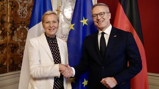 Karsten Wildberger und Anne Le Hénanff stehen nebeneinander und geben sich die Hand. Hinter ihnen sind die Flagge Frankreichs, Deutschlands und der Europäischen Union zu sehen.