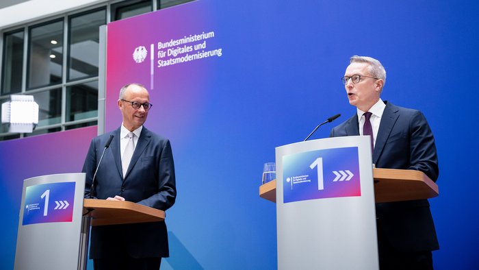Friedrich Merz und Karsten Wildberger stehen auf der Bühne vor der BMDS-Pressewand, Wildberger spricht ins Miktofon. 