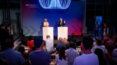 Blick auf die Bühne, auf der ein Mann und eine Frau stehen. Der Mann hält ein Mikrofon. Im Vordergrund das Publikum. 