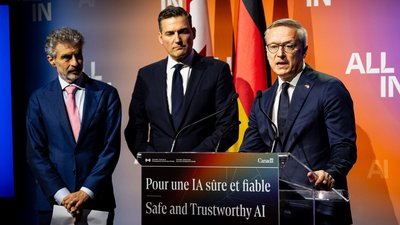 Drei Männer im Anzug stehen auf der Bühne vor der ALL IN-Wand. Einer redet am Rednerpult. 