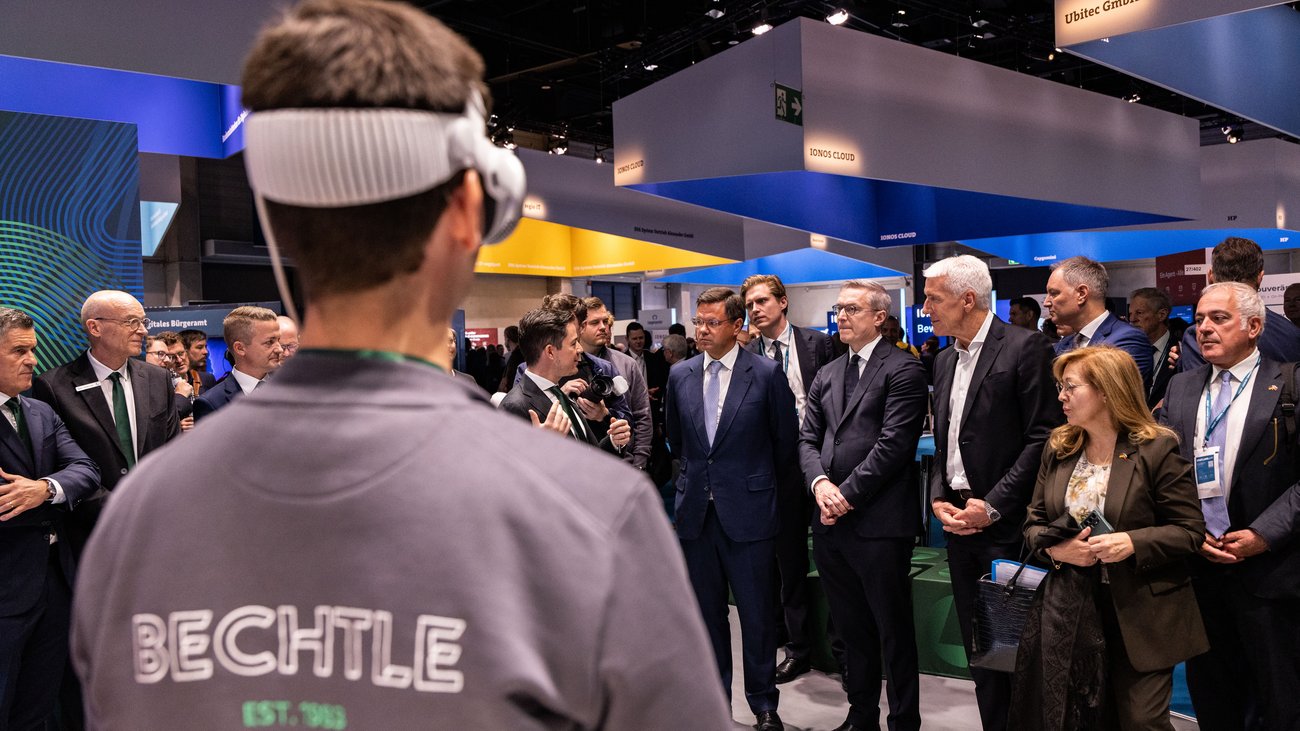 Ein Mann mit VR-Brille von hinten, vor ihm sprechen Messebesucher in ein Mikrofon