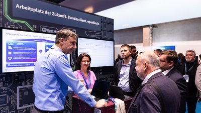 Situation an einer Messestation: An einem Stehtisch stehen 4 Menschen, im Hintergrund liest man "Arbeitsplatz der Zukunft. Bundesclient. openDesk.". Man sieht 2 Monitore. 