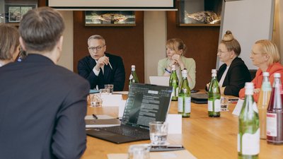 Mehrere Personen sitzen an einem Konferenztisch in einem Besprechungsraum. Auf dem Tisch stehen Wasserflaschen, Gläser und ein Laptop.